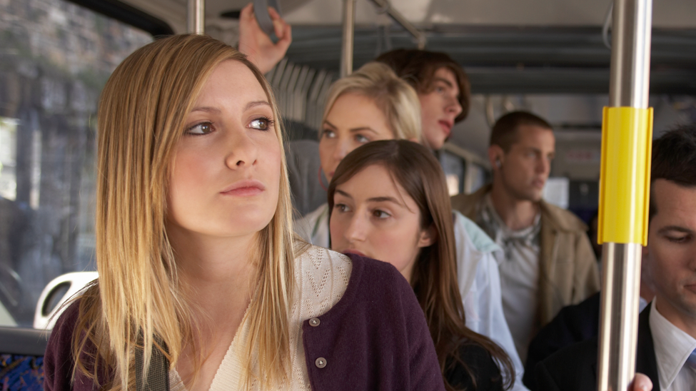 A woman with blonde hair and a purple cardigan stood on a bus, holding a metal pole