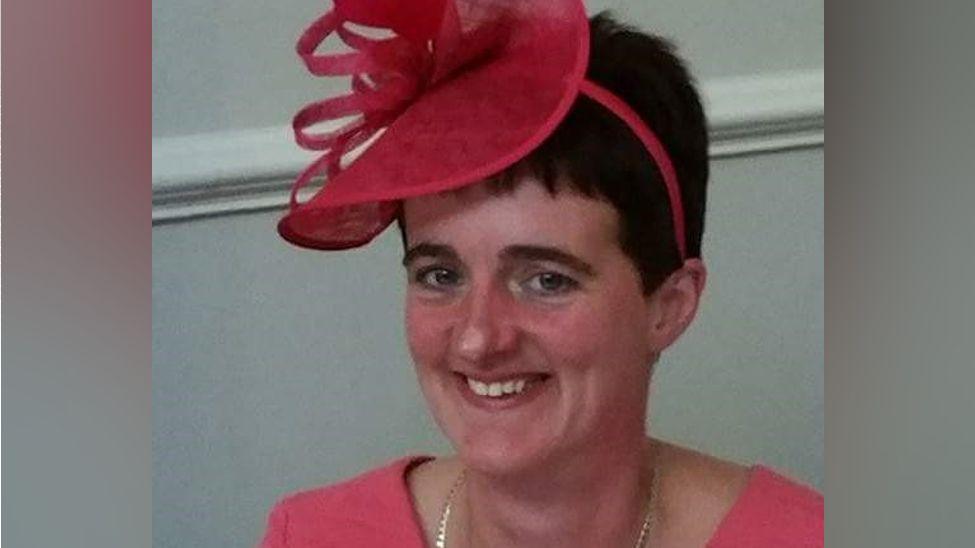 A woman wearing a pink fascinator and pink outfit smiles at the camera.