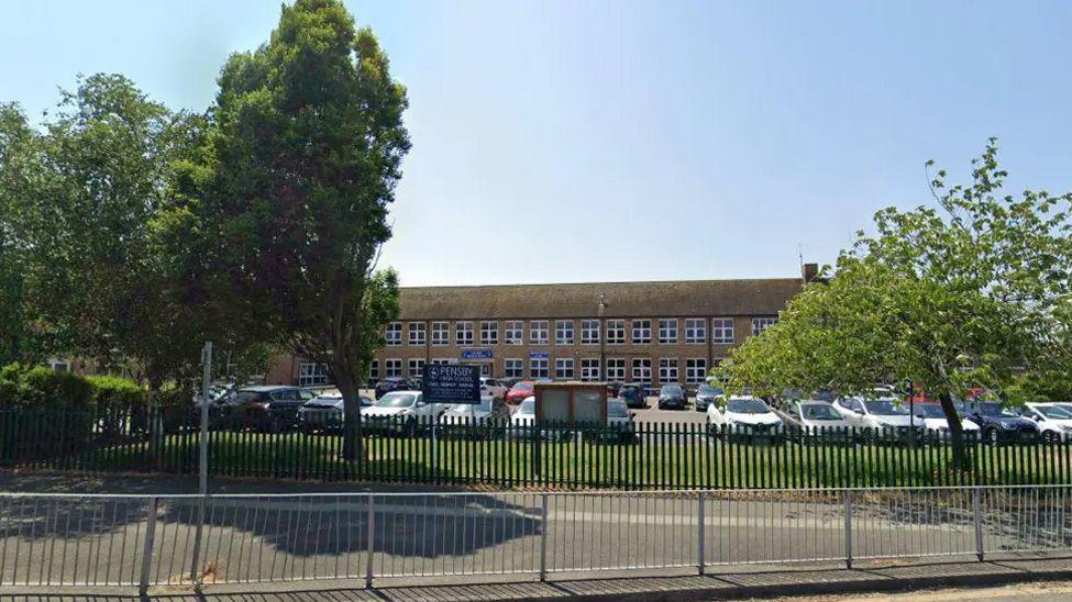 A two storey building is set behind trees and a metal fence, with a is a large car park in front almost full of cars.