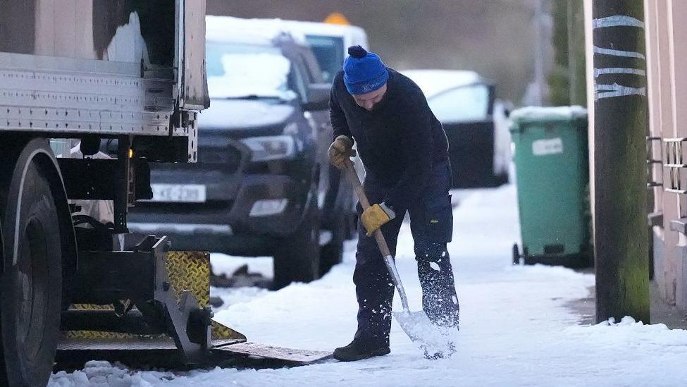 A man wearing a cap and heavy clothing uses a shovel to clear snow from a pavement in a street where cars and a lorry are parked.