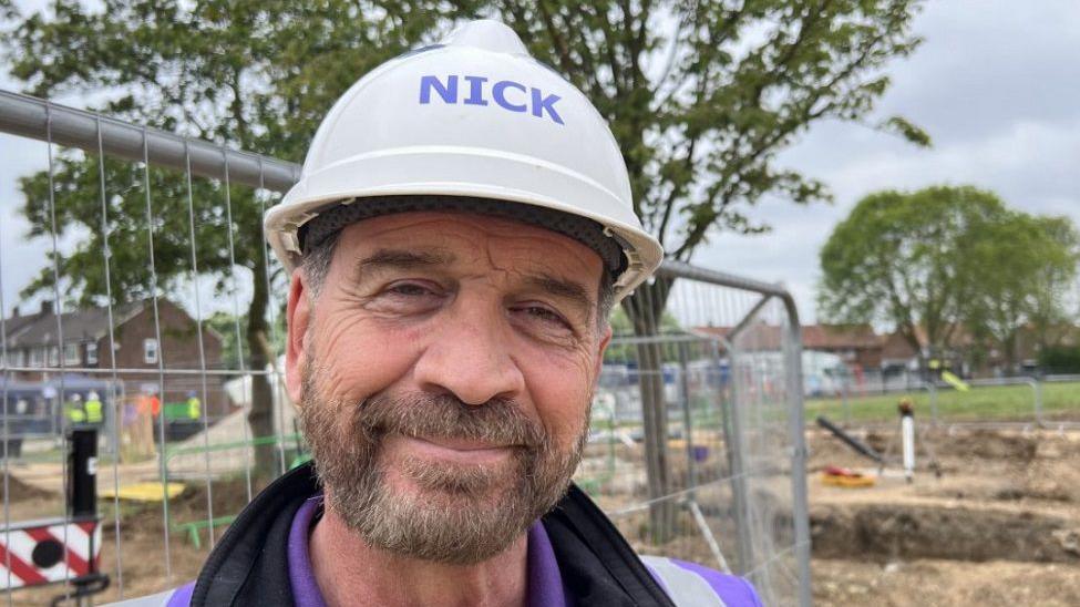 DIY SOS presenter Nick Knowles wearing a hard hat with the word: "Nick" on it and a purple hi-vis jacket. He is smiling with a brown beard and is stood in front of metal railings and work going on in some grass in the background.
