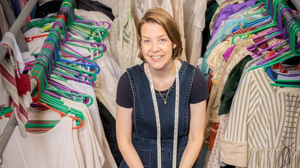 A woman with short brown hair smiling into the camera. She's surrounded by rails of clothes and costumes. She is wearing a denim dress and has a measuring tape around her neck.