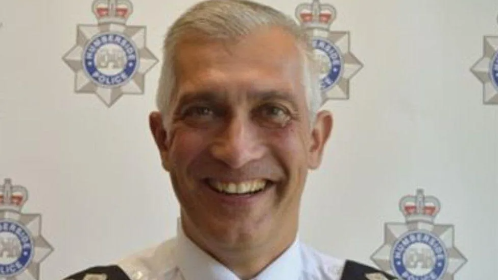 Paul Anderson has short cropped grey hair and is wearing a police uniform and is stood against a backdrop that shows logos of the Humberside force