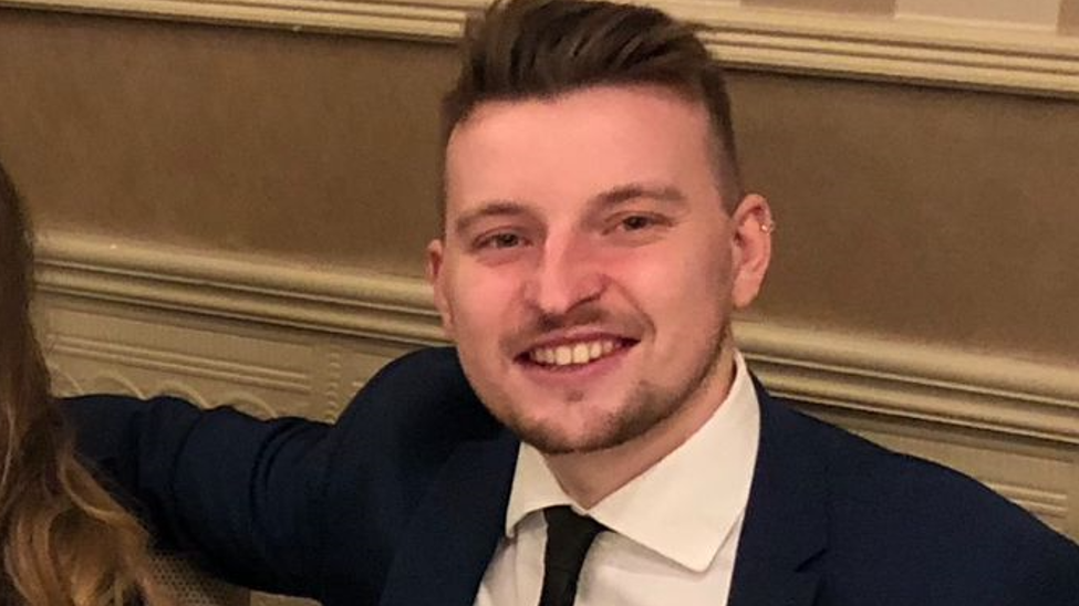 A smiling man with brown hair and a small beard in a dark suit, white shirt and dark tie