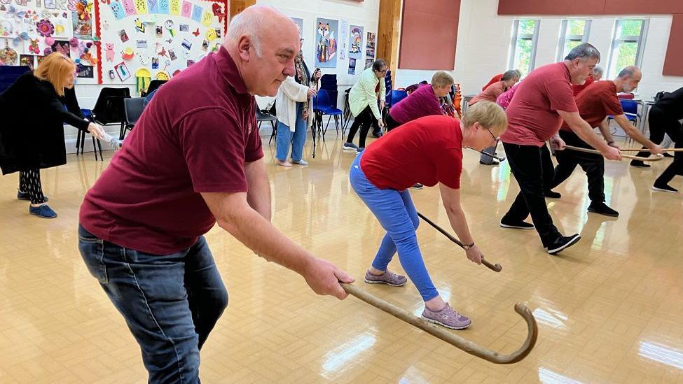 A group of older people learning Tai Chi with walking sticks in a community hall