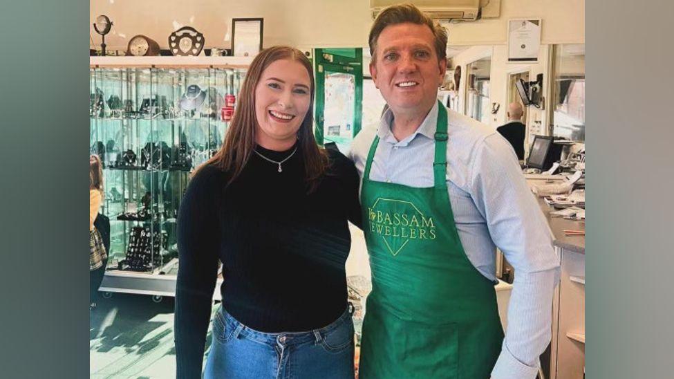 A woman and a man stand next to each other in a jewellery shop. The woman is wearing a black top, jeans and a silver necklace with a small pendant. The man is wearing a green apron over a white shirt. Behind them, there are glass cabinets containing jewellery on the left and a desk on the right.