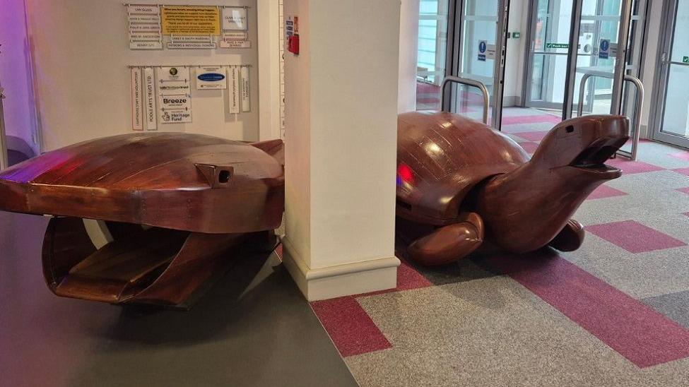 The wooden turtle and whale set up inside a building next to a pillar. 