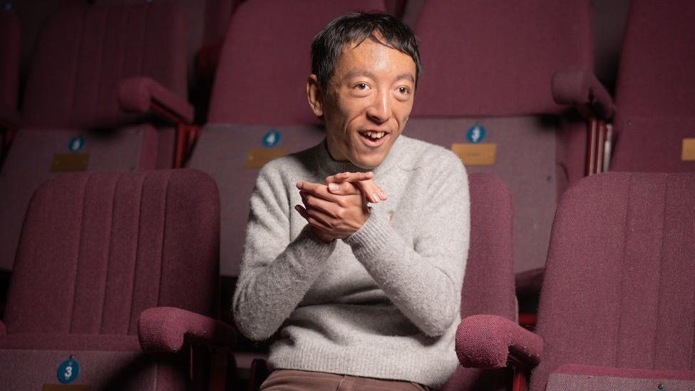 A man with dark hair sitting on a purple theatre seat. He is smiling with his hands clasped together in front of his chest.