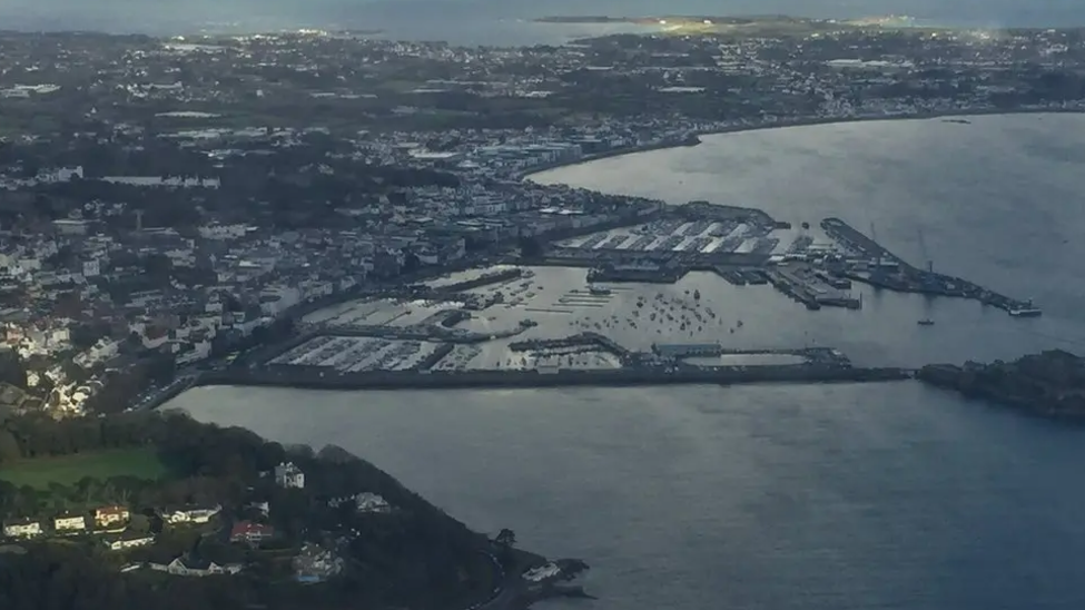 An aerial shot of Guernsey. There are houses on land on the left and the sea on the right.  