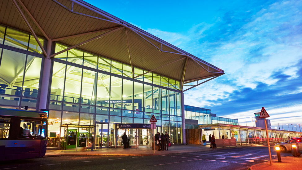 A night-time shot of Bristol Airport's terminal. Silhouettes of passengers are visible in the background. The terminal is brightly lit and the sky is dark blue  