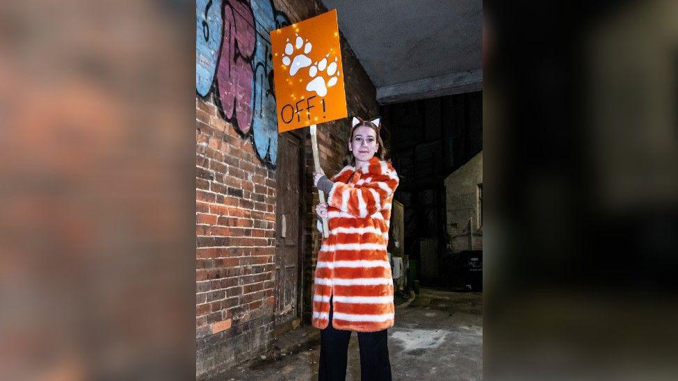 A woman wearing an orange and white stripy fluffy coat and a pair of fox ears, holding up a sign saying "paws off". She's outside and there's a graffitied brick wall behind her.