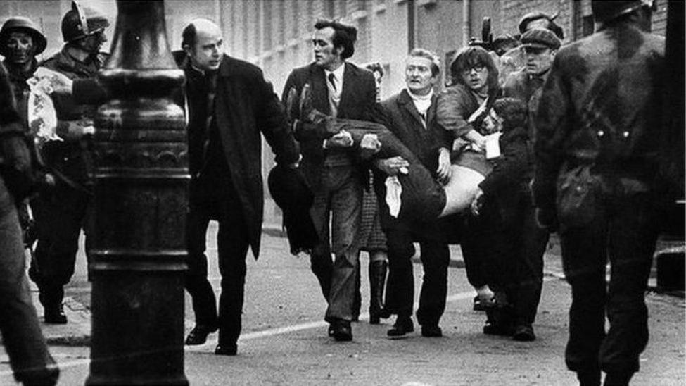 A number of men carry a younger man who has been shot on Bloody Sunday. A priest can be seen waving a white hankie, while a number of armed soldiers are in the backgorund