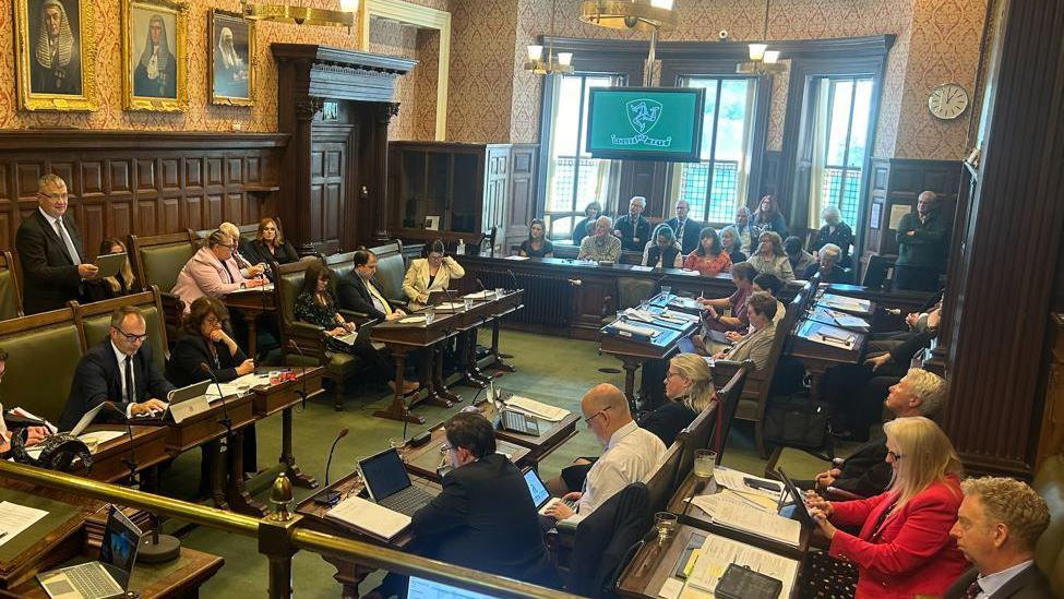 Members of the House of Keys sitting on benches in the chamber, with members of the public watching on from the public gallery.