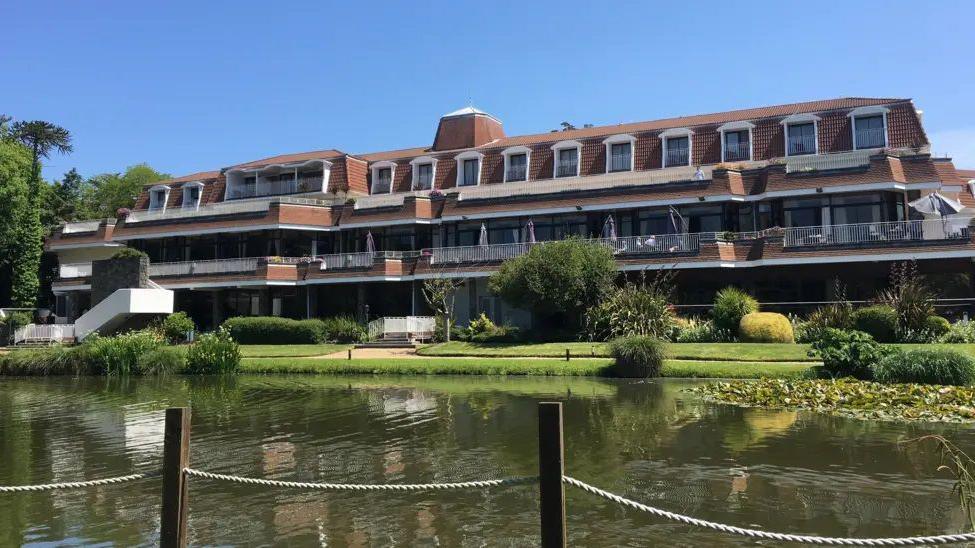 St Pierre Park is a red brick hotel with balconies that has planting and a lake in front of it. It is a blue sky day and there is a post and chain fence in the foreground.