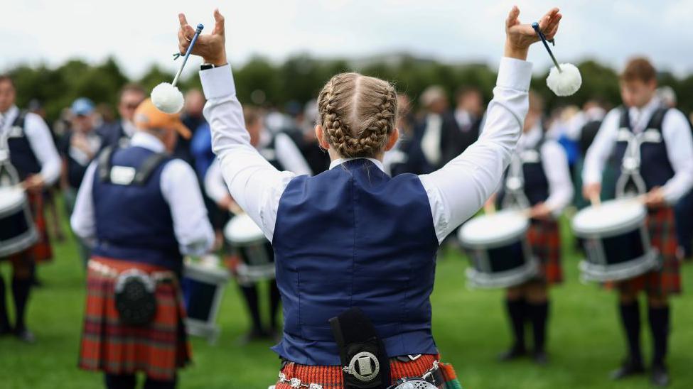 Back of a female tenor drummers head. She is wearing a navy waistcoat and a red kilt. She is twirling tenor sticks in the air.