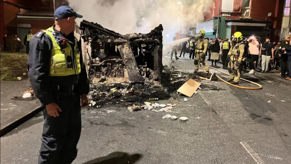 A burnt-out bus lies in the street with crowds and emergency services looking on 