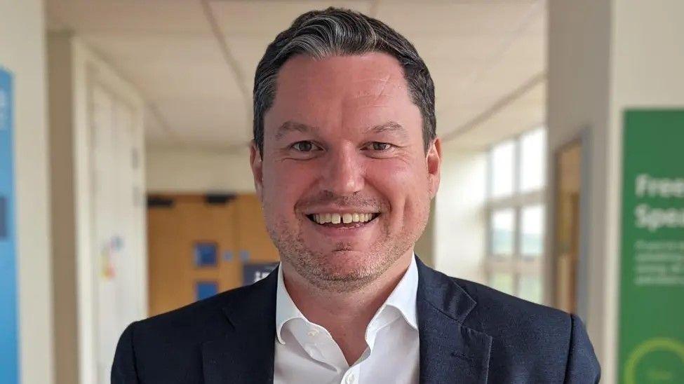 A picture of Kevin McNamara in a corridor, he is wearing a navy blazer and a white shirt, and smiling widely.