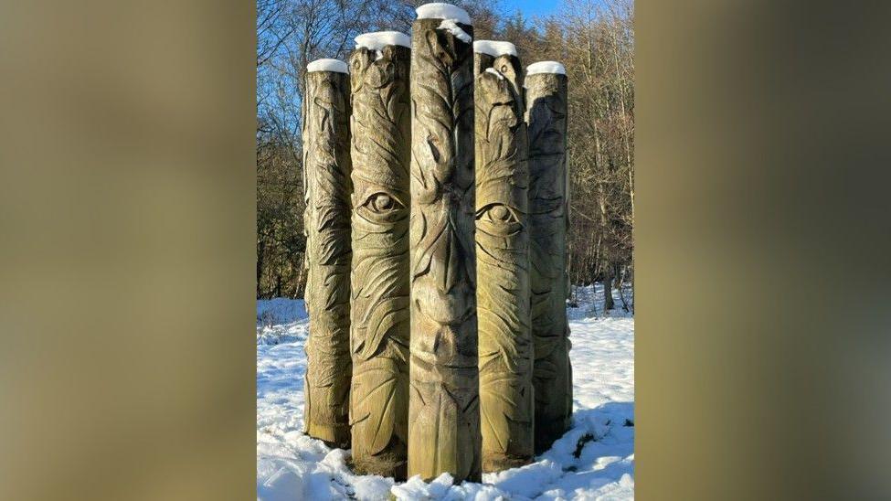 Five wooden poles standing in the snow. Each has carvings which when photographed together show the face of the Green Man. Trees can be seen in the background.