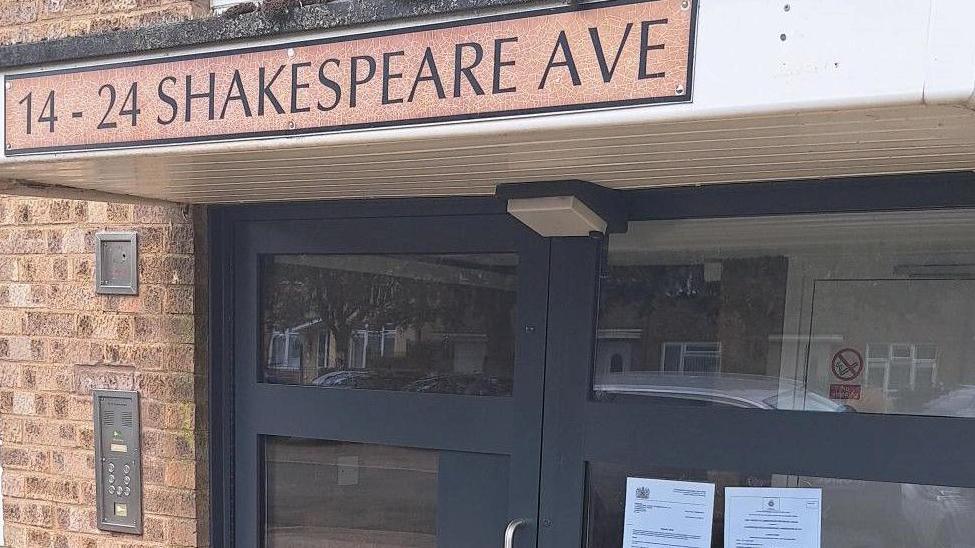 A glass-panelled front door to a block of flats, with a sign above the door reading: "14-24 Shakespeare Ave."