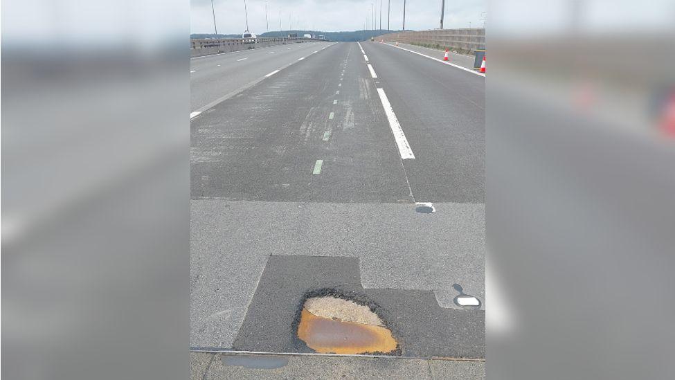 The M5 motorway on the Avonmouth Bridge. At the bottom of the picture there is a large pothole that has formed on the tarmac, with darker patches of repair work previously done around it. 