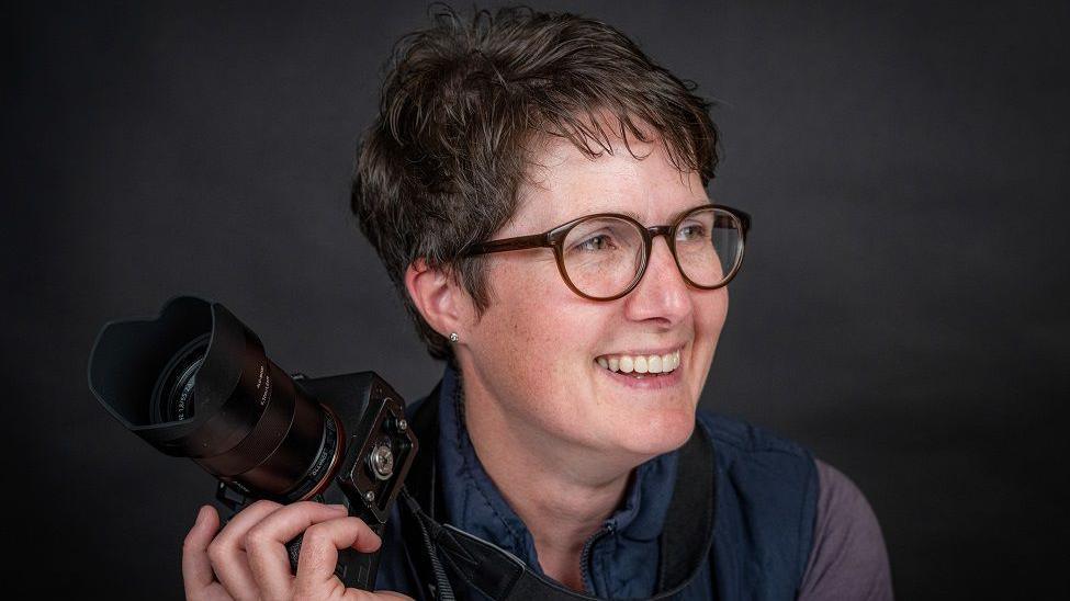 A woman with brown short hair and brown glasses smiling whilst looking to the right. She is holding a large camera.