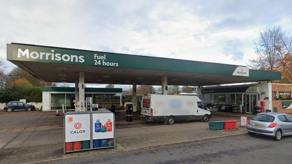 A Morrisons fuel station. Key features include: A green canopy with the Morrisons logo and the words "Fuel 24 hours". Multiple fuel pumps with several cars parked nearby. An attached convenience store. Surrounding autumnal trees and a paved road leading into the station.