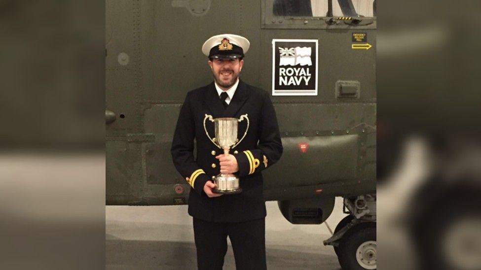 Steve Irwin standing in front of a helicopter in Royal Navy uniform holding a large silver trophy and smiling