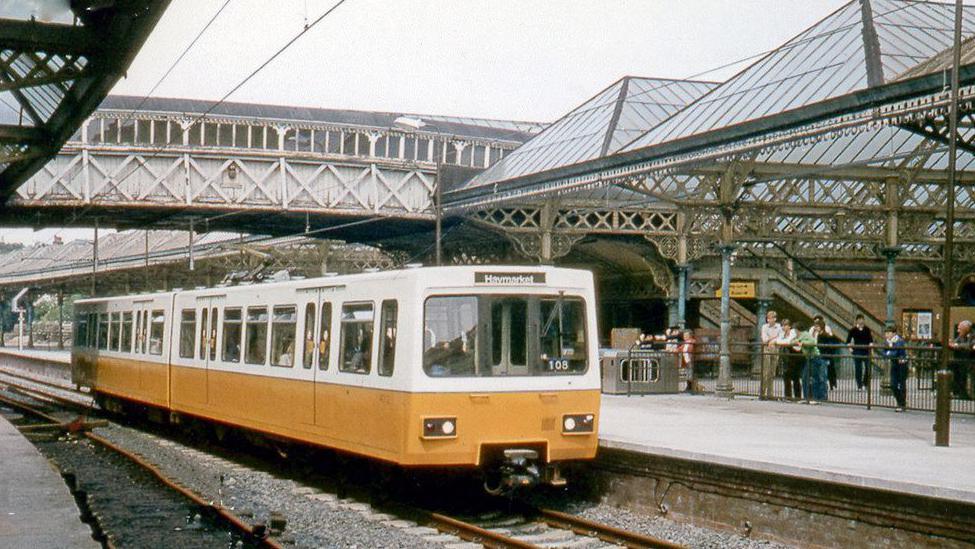 A photo from 1980 of one of the original Tyne and Wear Metro trains. It is in yellow and white livery, with the destination marked as Haymarket, and is standing a Tynemouth Metro Station, with people looking at it over a railing.