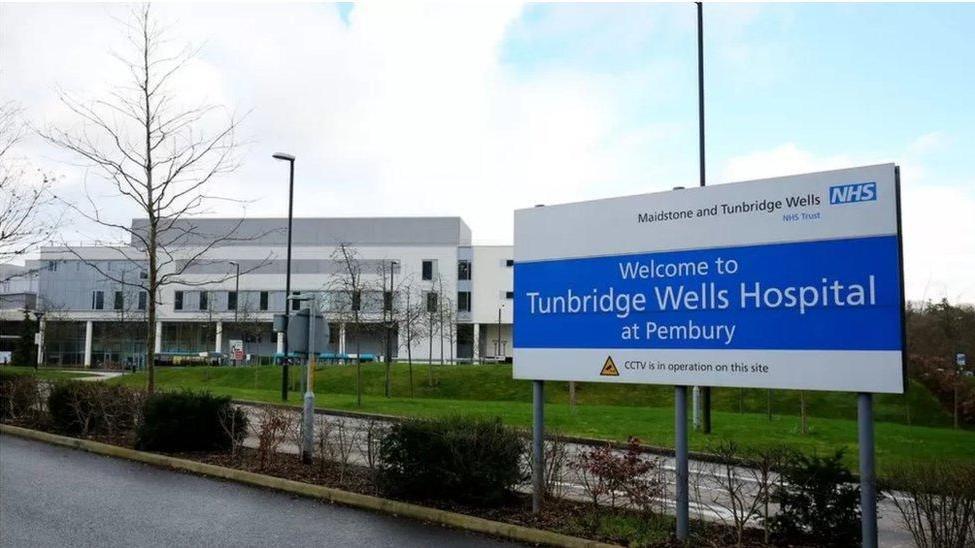 A sign outside Tunbridge Wells Hospital. There is a hospital building in the background, as well as a green grass and bushes. 