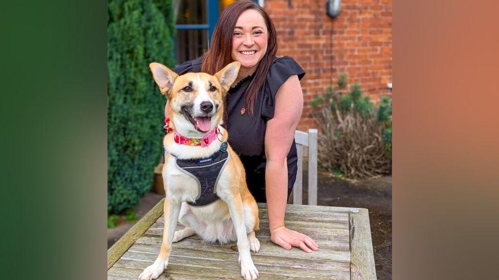 A woman stands behind a dog in a pub garden. The dog has light brown and white fur and is sitting on a wooden table. The woman has straight brown hair, and she is wearing a black top with a small red poppy brooch. 