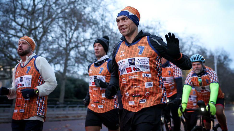 Former rugby player Kevin Sinfield runs with a team of supporters as he waves to crowds while on one of his previous fundraising challenges. The runners wear orange and blue matching vests and headbands and are joined by a man on a bike.