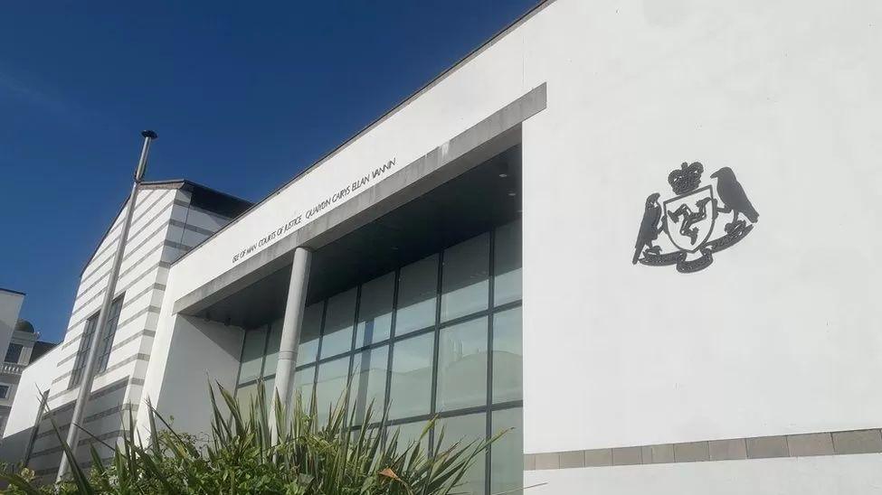 The exterior of the grand white Isle of Man Courts of Justice building, featuring the Manx government's crest in black on the wall on the right hand side.