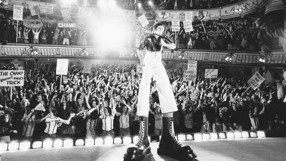 Elton John wearing a cap and very long white trousers with giant black Dr Martens boots on his feet.  He is singing on a stage with people behind him swinging their arms to the music.  There is a row of footlights in front of the crowd.  The picture is in black and white from the 1970s.