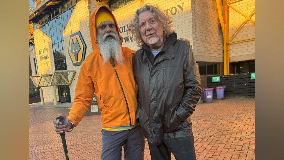 Two men standing in front of a football stadium. One is wearing an orange jacket and has a yellow turban. The other is wearing a black leather jacket.