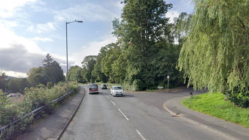 A7 Abbotsford Road through Galashiels. It is a 30mph road with one lane of cars going in each direction. A junction is seen in the picture with a 20mph speed limit sign. Pavements are at the side of the road as well as large trees and grass. 