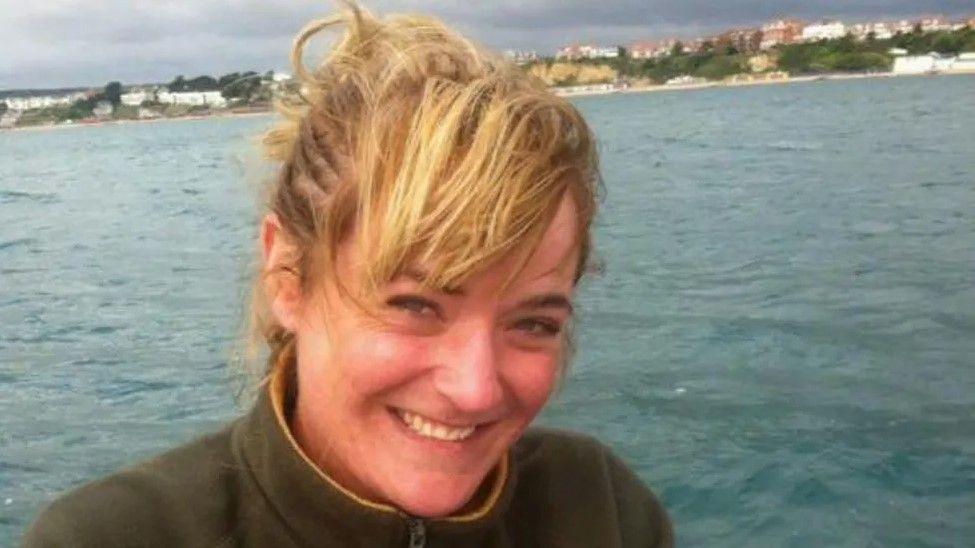 A woman with blonde air and a brown sweater with a yellow rim smiles at the camera, with the sea in the background