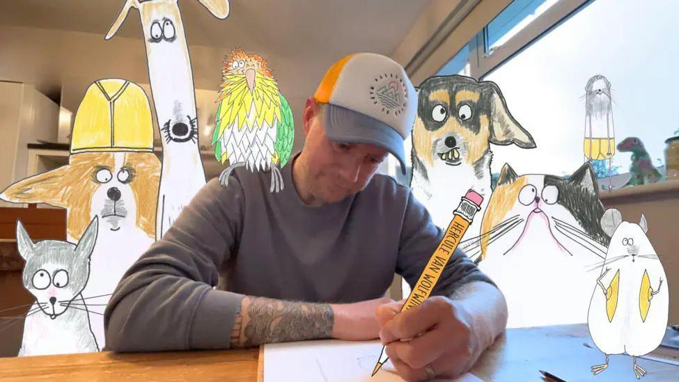 Phil Heckels sat wearing a cap drawing holding a large cut out pen with large cardboard cutouts of some of his artwork surrounding him.
