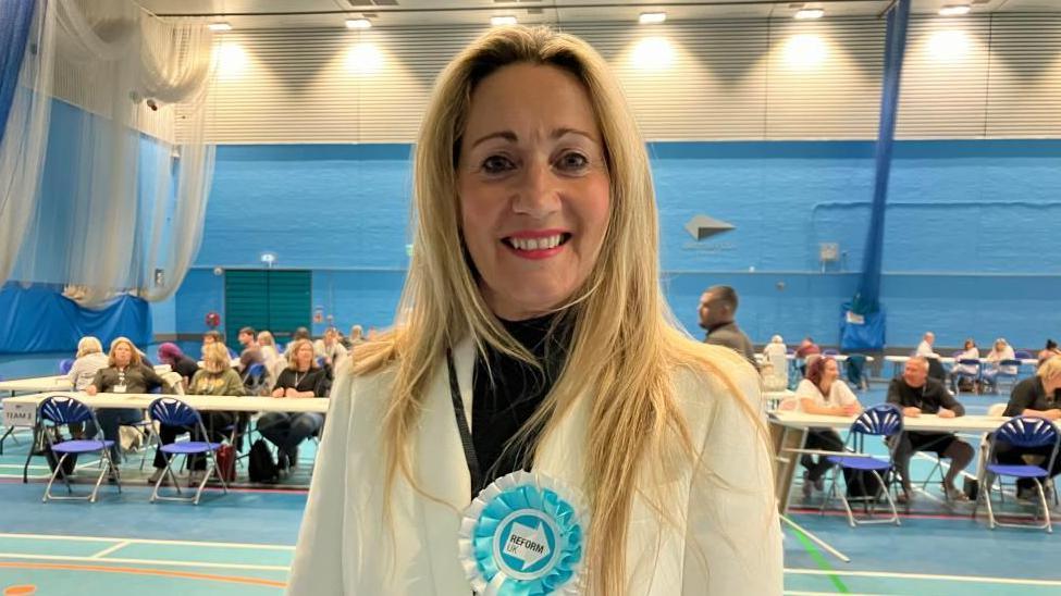 A woman is standing in a gym hall which is being used for counting in elections. She has long blonde hair, she is smiling and is wearing a white jacket with black roll neck underneath. On the jacket is a rosette which is light blue and says Reform on it. 