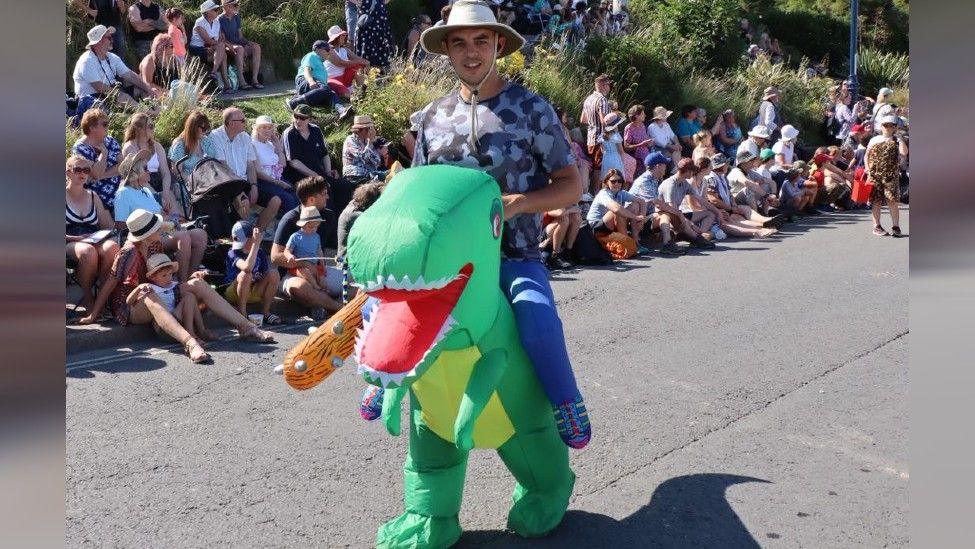 A man dressed in a dinosaur costume which makes it look like he is sat on the back of a green dinosaur.