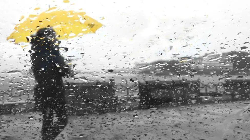 A person wearing a black coat and trousers holding a yellow umbrella. The foreground of the picture is covered in rain as if it is being viewed through a windscreen or window.