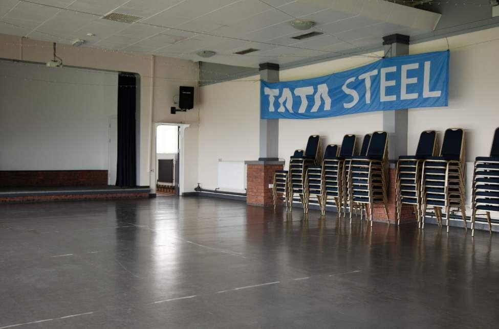 Clwb cymdeithasol gwag gyda phentwr o gadeiriau a baner las gyda'r geiriau 'Tata Steel' ar y wal