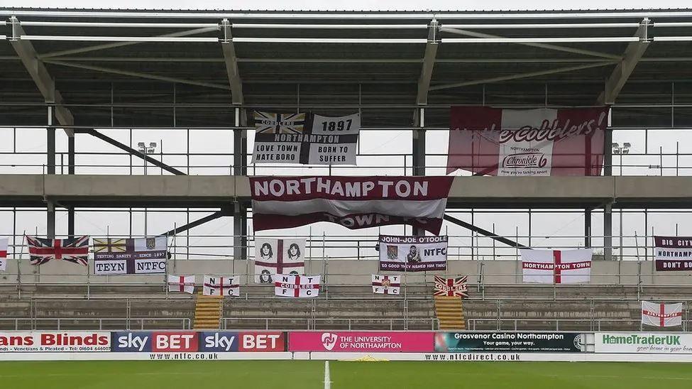 A half-built shell of a football stand draped in supporter flags with the pitch in the foreground.