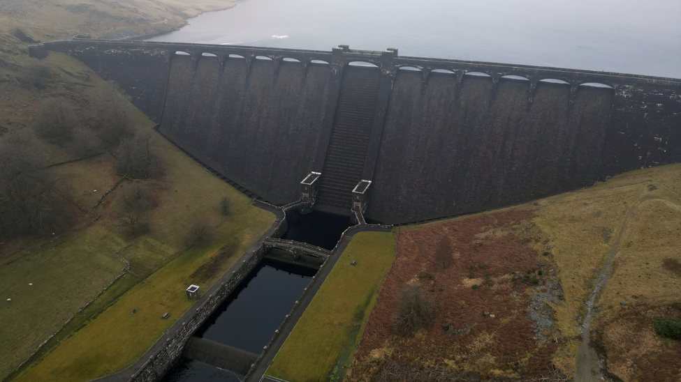 An aerial photo of the imposing Claerwen Dam in mid Wales. The dam is made from grey stone and has arches along the top. It is surrounded by barren misty hills.