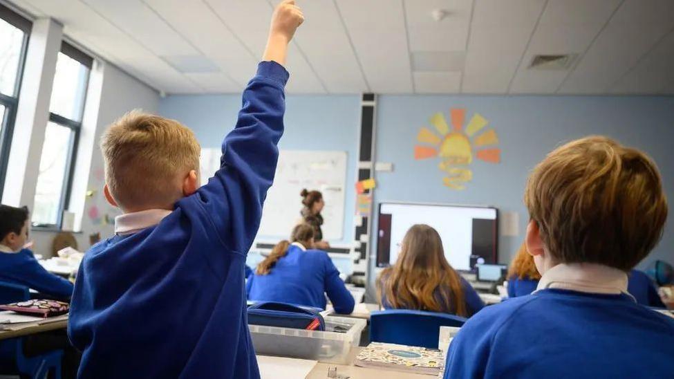 A child in a blue uniform raising his hand in a school classroom. He is sat next to other pupils but we can only see the backs of their heads as they all wear the same uniform of a blue jumper over a white shirt. 