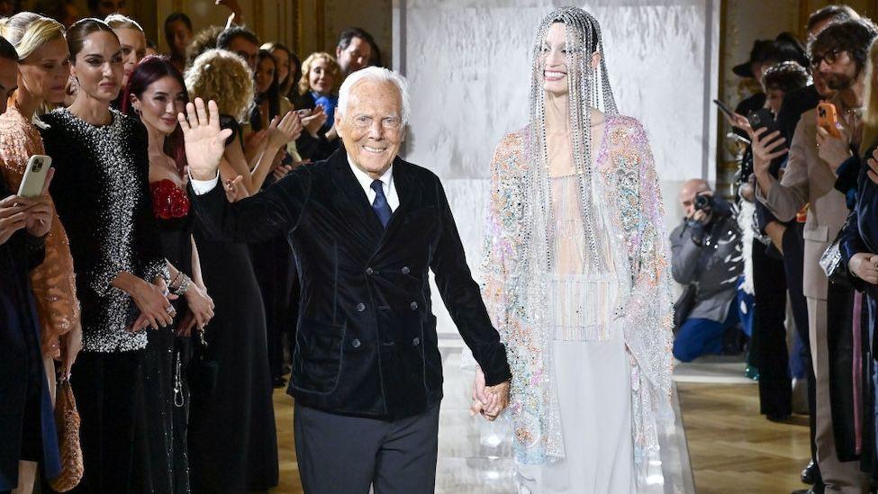 An elderly man in a velvet jacket holds the hand of a tall model, wearing beads across her face and a sheer, embellished dress.