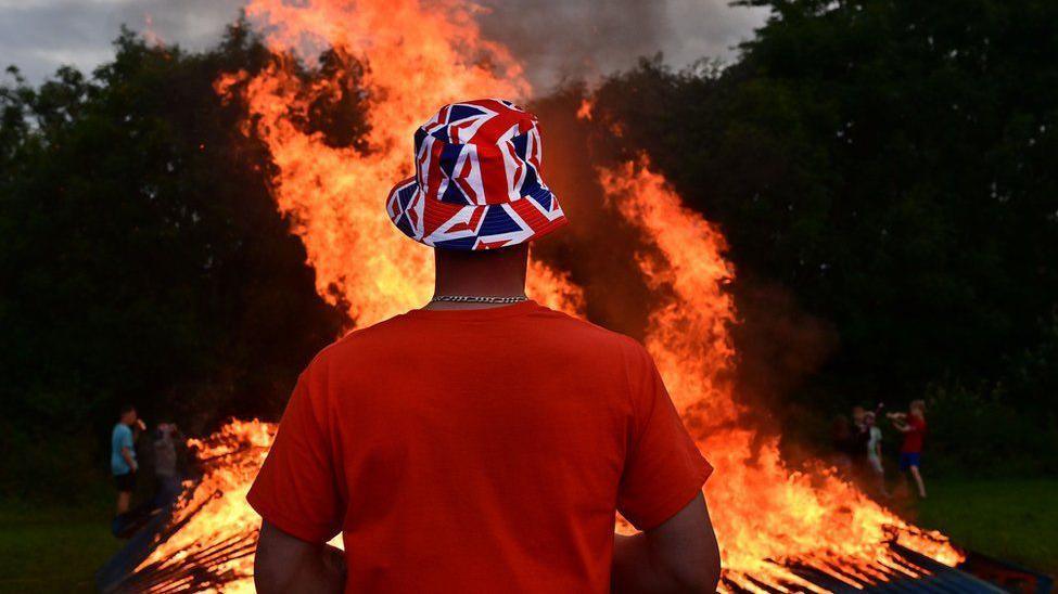 Man with UK flag hat in red, white and blue in front of a burning bonfire