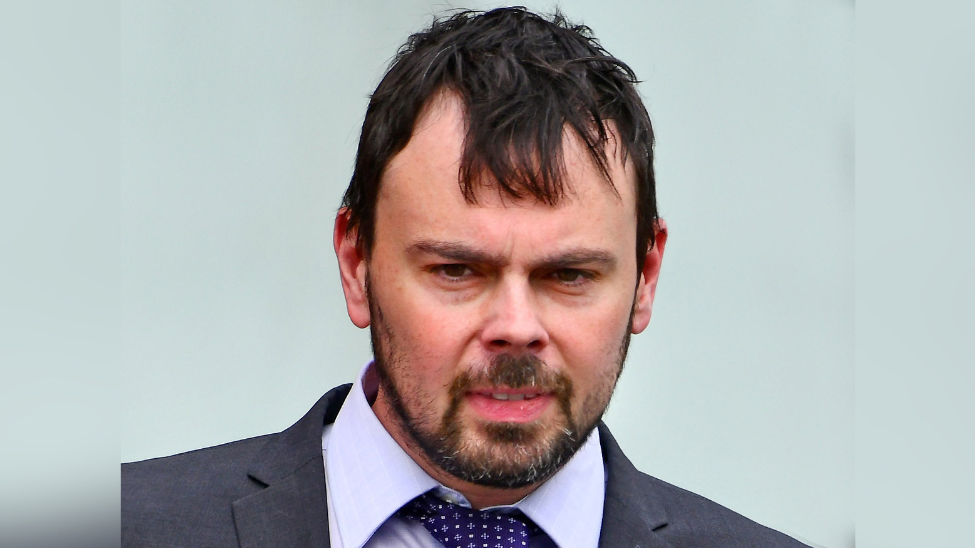 William Maher is wearing a grey suit with a tie. He has brown hair and is outside.