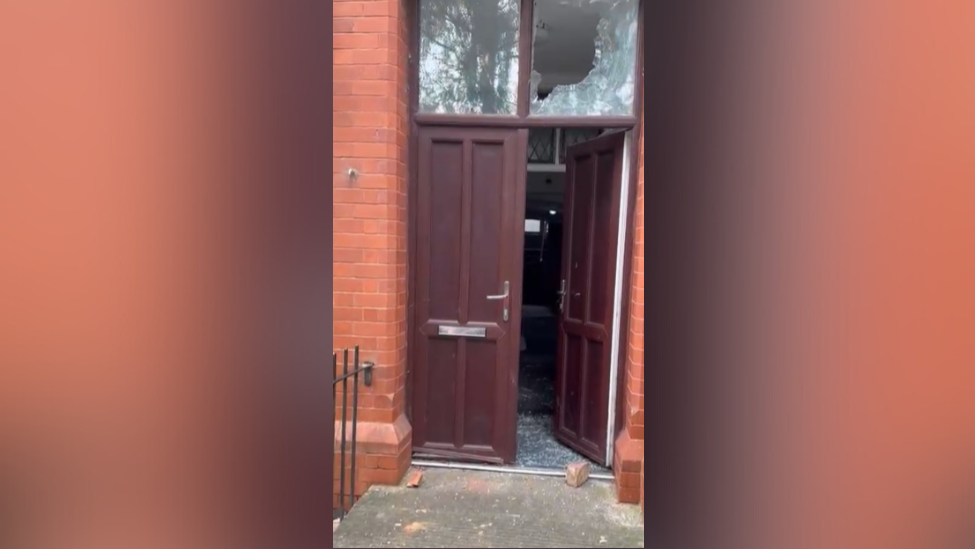 A door in a brick building has a smashed window above it and blocks of brick on the ground. 