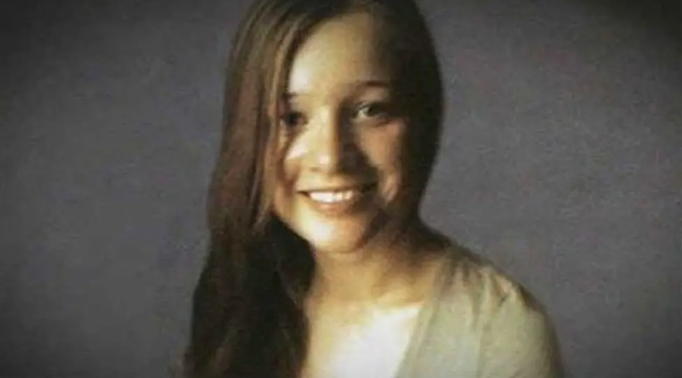 Angelika Kluk, a smiling young woman with long brown hair, wearing a scoop neck grey top, pictured against a grey background.