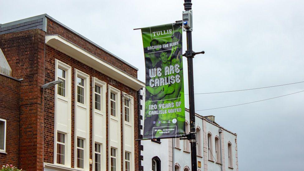 A green banner attached to a black lamppost with a photo of men hugging. The banner reads "Tullie- Backing the Blues- We Are Carlise- 120 years of Carlisle United." Carlisle is spelled with one l in the words 'We are Carlise'.